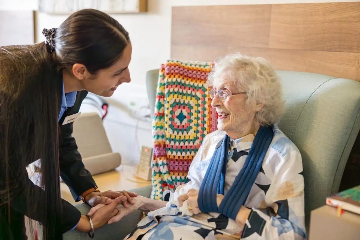 Aged care worker with brown hair talks to an older women knitting on a lounge chair